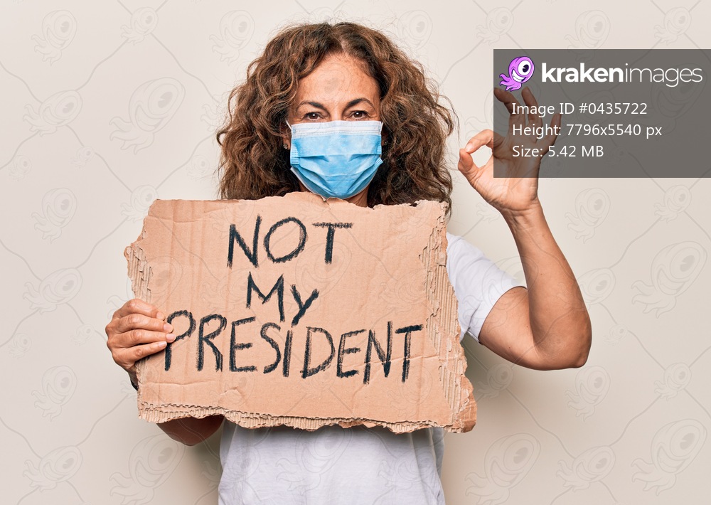 Middle age woman wearing coronavirus protection mask holding not my president protest cardboard doing ok sign with fingers, smiling friendly gesturing excellent symbol