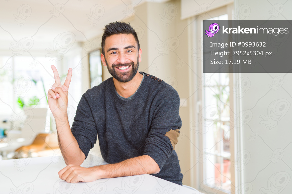Handsome hispanic man wearing casual sweater at home smiling with happy face winking at the camera doing victory sign. Number two.