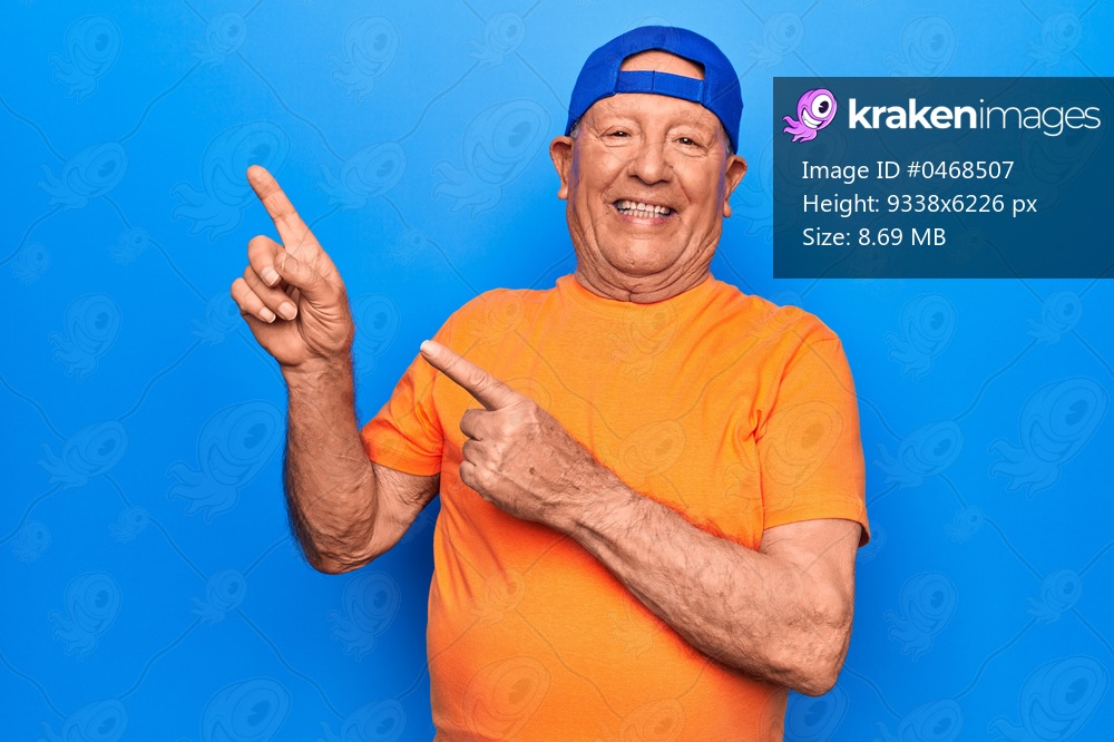 Senior handsome grey-haired man wearing casual t-shirt and cap over blue background smiling and looking at the camera pointing with two hands and fingers to the side.