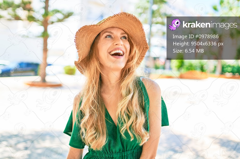 Young beautiful blonde woman on vacation wearing summer hat smiling happy. Standing with smile on face at street of city.