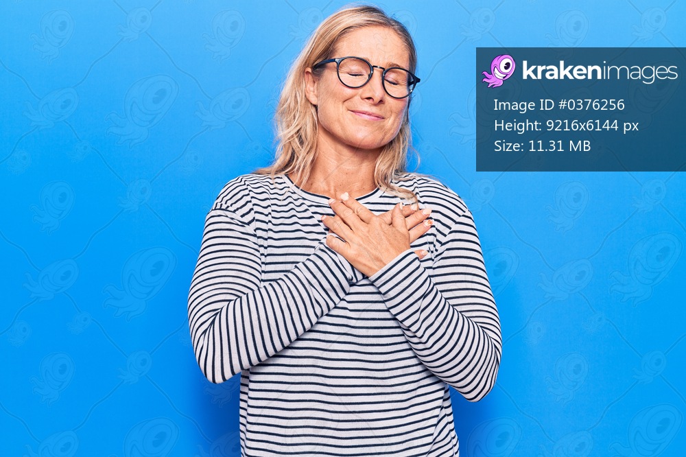 Middle age caucasian blonde woman wearing casual striped sweater and glasses smiling with hands on chest with closed eyes and grateful gesture on face. health concept. 