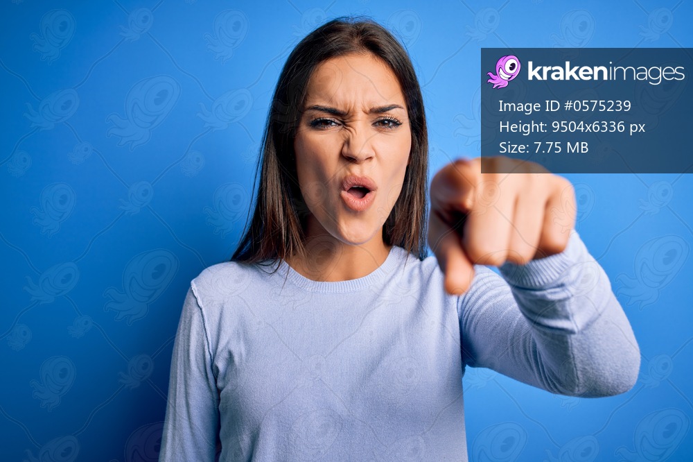 Young beautiful brunette woman wearing casual sweater standing over blue background pointing displeased and frustrated to the camera, angry and furious with you