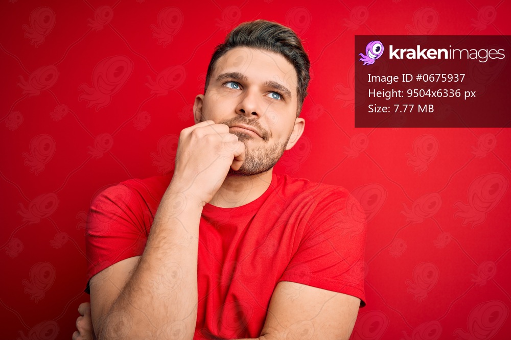 Young man with blue eyes wearing casual t-shirt over red isolated background with hand on chin thinking about question, pensive expression. Smiling with thoughtful face. Doubt concept.