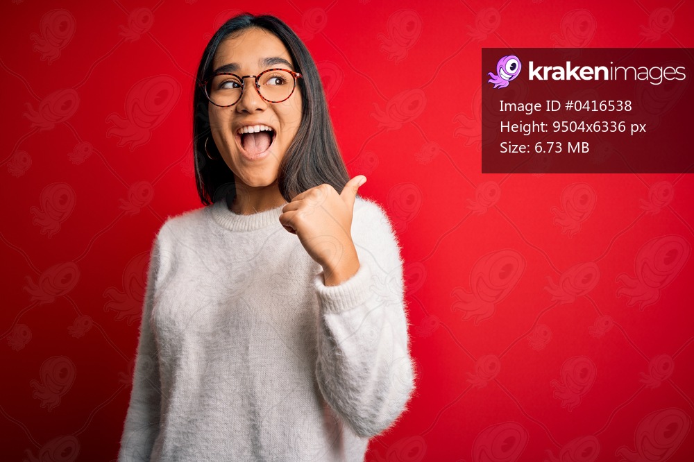 Young beautiful asian woman wearing casual sweater and glasses over red background smiling with happy face looking and pointing to the side with thumb up.