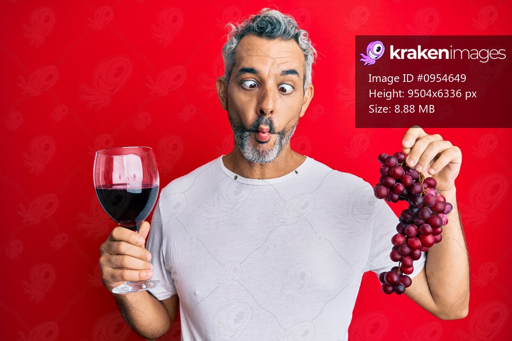 Middle age grey-haired man holding branch of fresh grapes and red wine making fish face with mouth and squinting eyes, crazy and comical. 