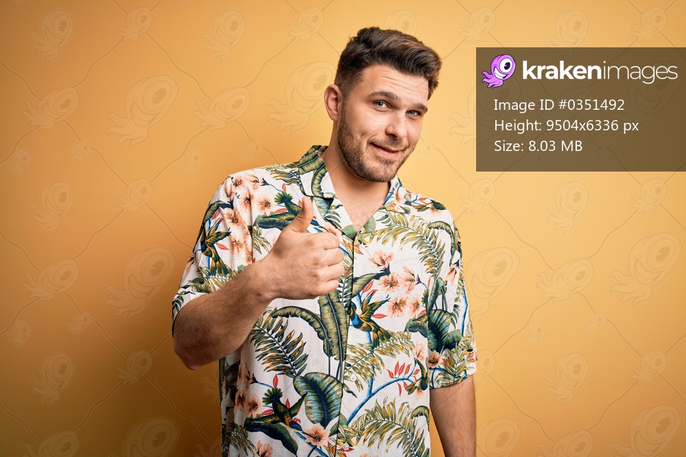 Young man with blue eyes on vacation wearing floral summer shirt over yellow background doing happy thumbs up gesture with hand. Approving expression looking at the camera showing success.