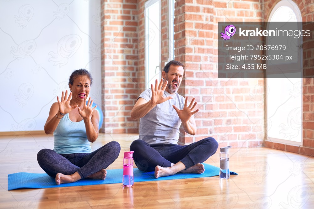 Middle age sporty couple sitting on mat doing stretching yoga exercise at gym afraid and terrified with fear expression stop gesture with hands, shouting in shock. Panic concept.