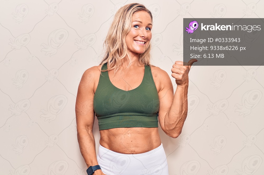 Middle age caucasian blonde woman wearing sportswear smiling with happy face looking and pointing to the side with thumb up. 