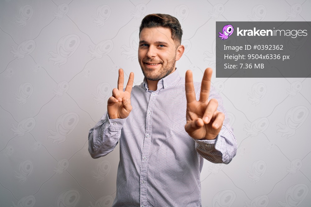 Young business man with blue eyes standing over isolated background smiling looking to the camera showing fingers doing victory sign. Number two.