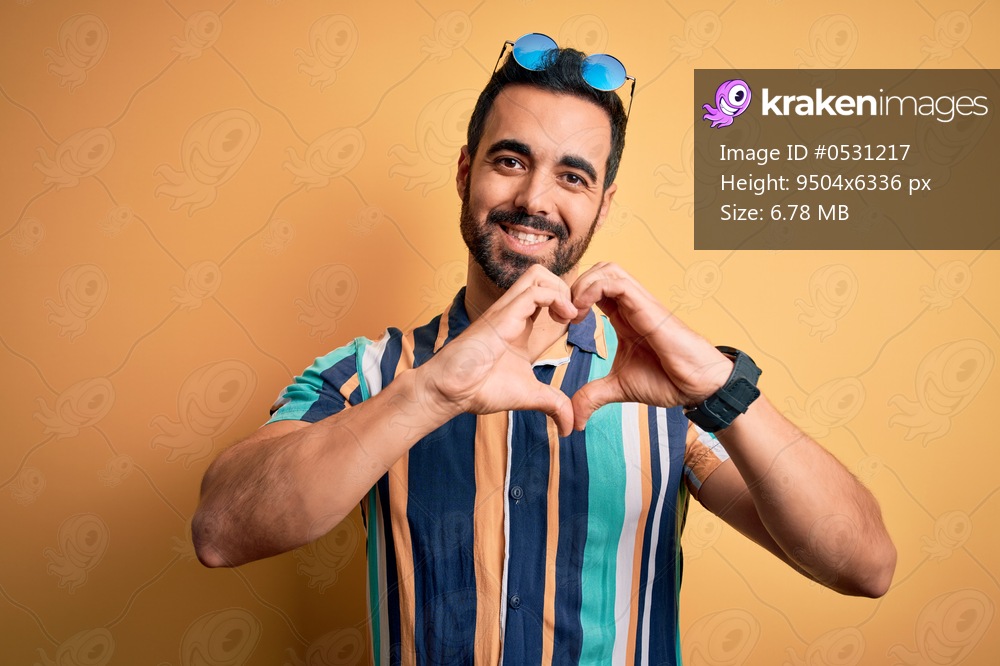 Handsome tourist man with beard on vacation wearing summer striped shirt and sunglasses smiling in love showing heart symbol and shape with hands. Romantic concept.