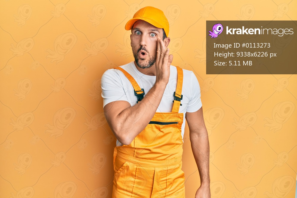 Young handsome man wearing handyman uniform over yellow background hand on mouth telling secret rumor, whispering malicious talk conversation 