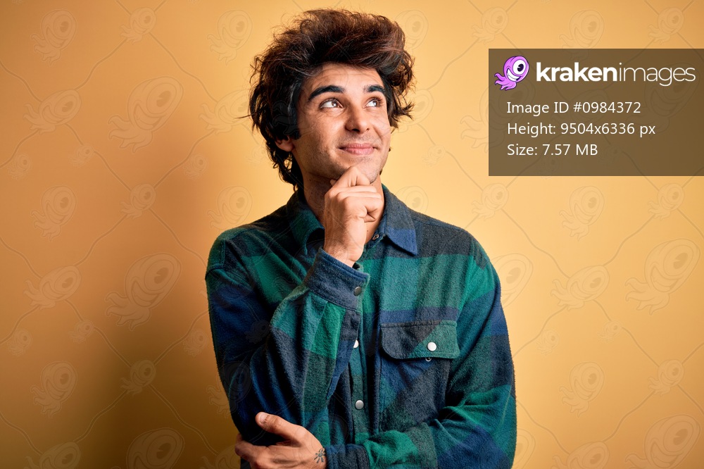 Young handsome man wearing casual shirt standing over isolated yellow background with hand on chin thinking about question, pensive expression. Smiling and thoughtful face. Doubt concept.