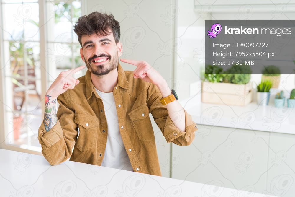 Young man wearing casual jacket sitting on white table smiling confident showing and pointing with fingers teeth and mouth. Health concept.