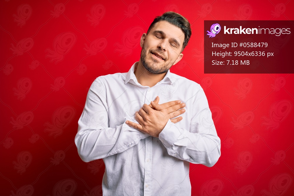 Young business man with blue eyes wearing elegant shirt standing over red isolated background smiling with hands on chest with closed eyes and grateful gesture on face. Health concept.