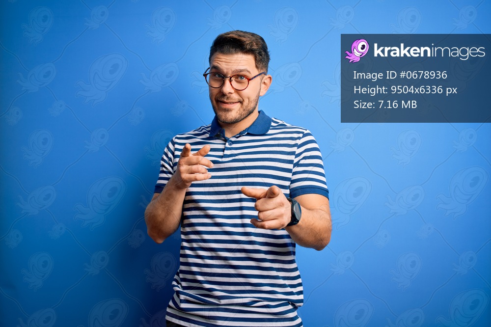 Young man with blue eyes wearing glasses and casual striped t-shirt over blue background pointing fingers to camera with happy and funny face. Good energy and vibes.