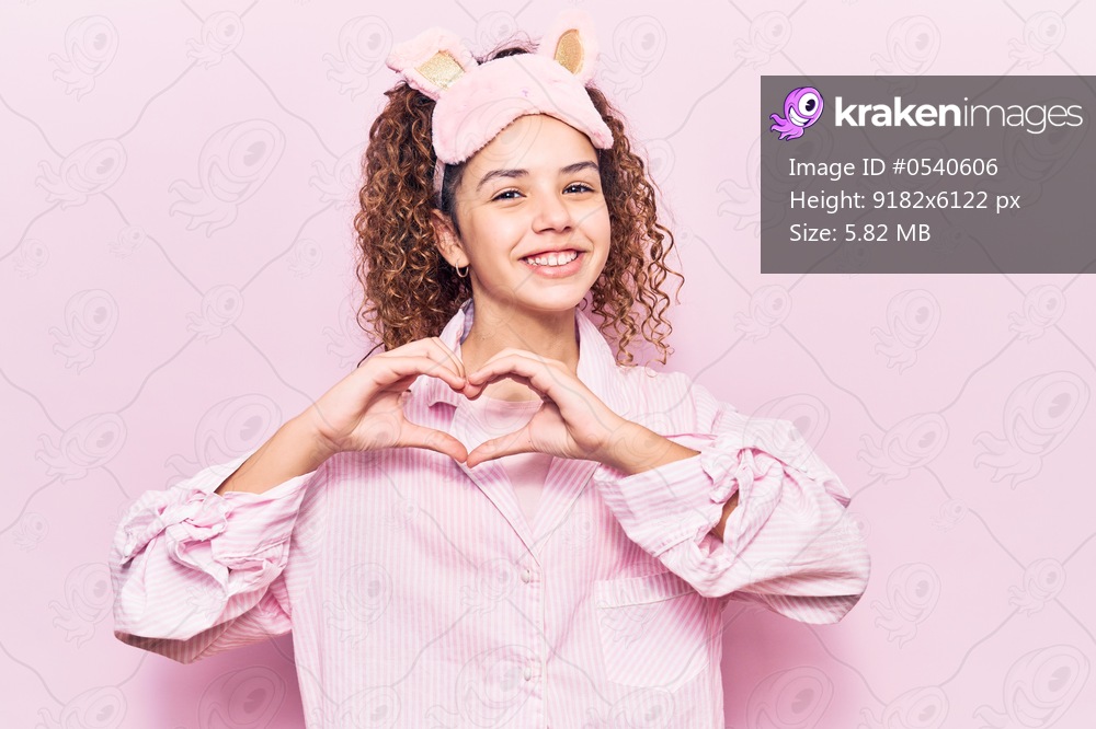 Beautiful kid girl with curly hair wearing sleep mask and pajamas smiling in love showing heart symbol and shape with hands. romantic concept. 