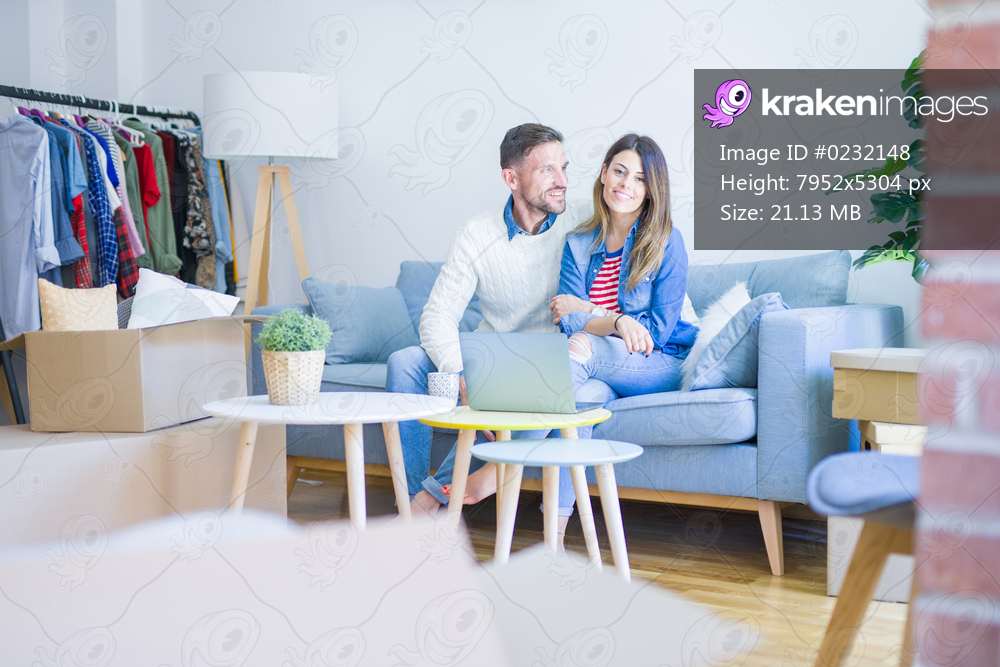 Young beautiful couple sitting on the sofa drinking cup of coffee using laptop at new home around cardboard boxes