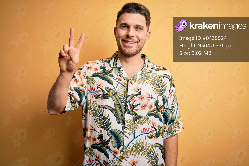 Young man with blue eyes on vacation wearing floral summer shirt over yellow background showing and pointing up with fingers number two while smiling confident and happy.