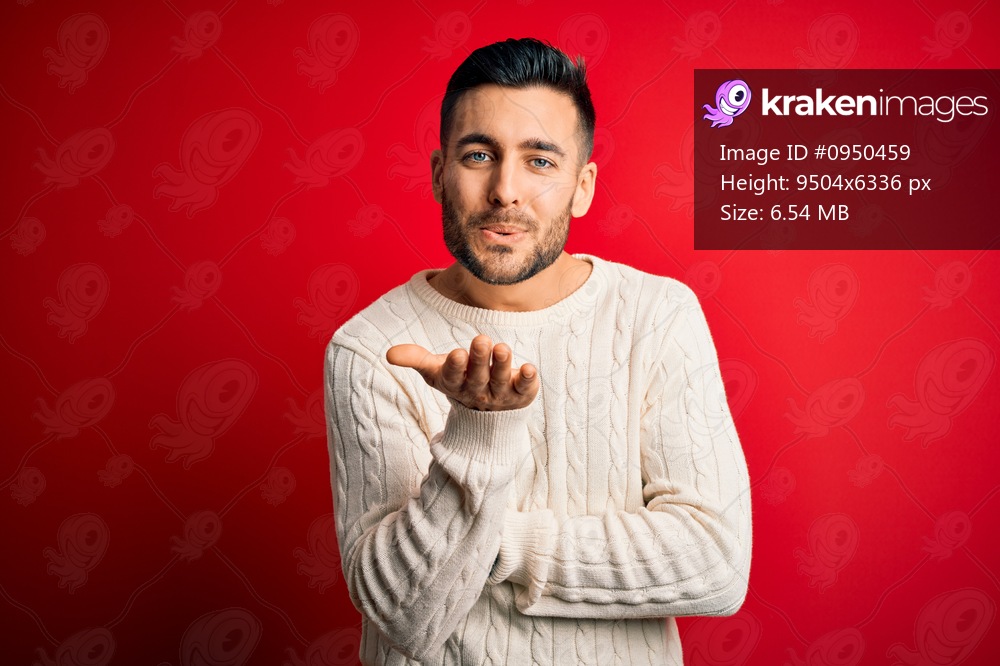Young handsome man wearing casual white sweater standing over isolated red background looking at the camera blowing a kiss with hand on air being lovely and sexy. Love expression.