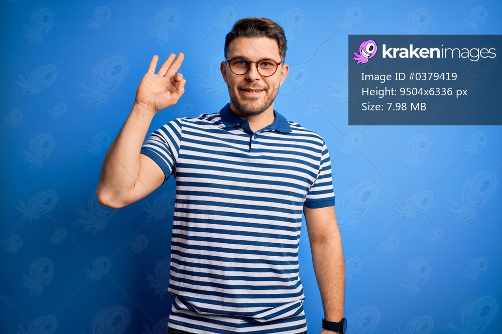 Young man with blue eyes wearing glasses and casual striped t-shirt over blue background smiling positive doing ok sign with hand and fingers. Successful expression.