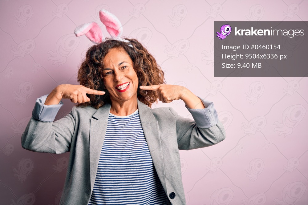 Middle age beautiful woman wearing bunny ears standing over isolated pink background smiling cheerful showing and pointing with fingers teeth and mouth. Dental health concept.