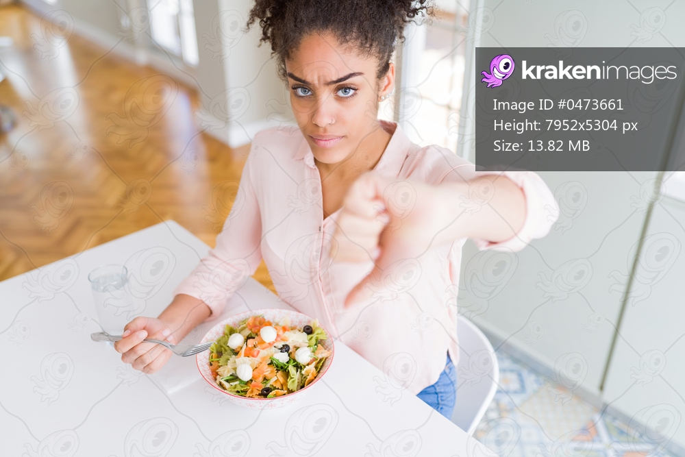 Young african american woman eating healthy pasta salad with angry face, negative sign showing dislike with thumbs down, rejection concept