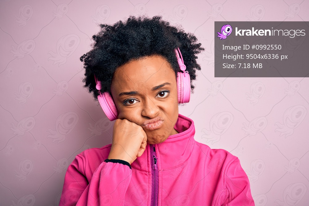 Young African American afro woman with curly hair listening to music using pink headphones thinking looking tired and bored with depression problems with crossed arms.