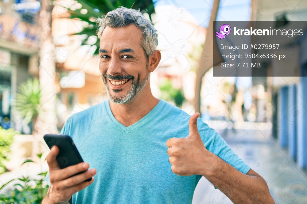 Middle age grey-haired man using smartphone at street of city smiling happy and positive, thumb up doing excellent and approval sign 