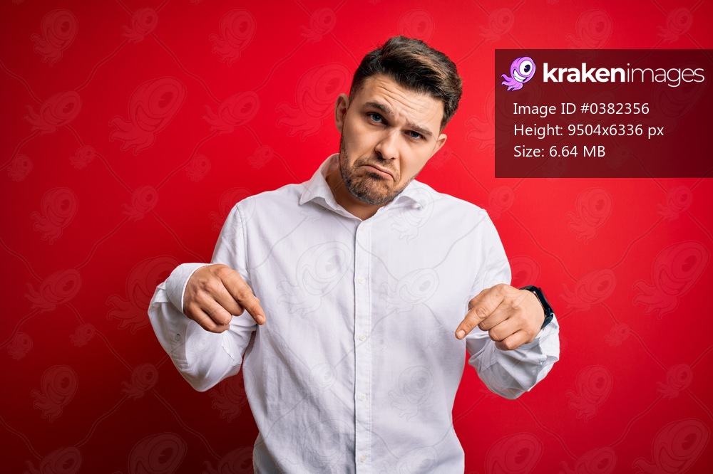 Young business man with blue eyes wearing elegant shirt standing over red isolated background Pointing down looking sad and upset, indicating direction with fingers, unhappy and depressed.