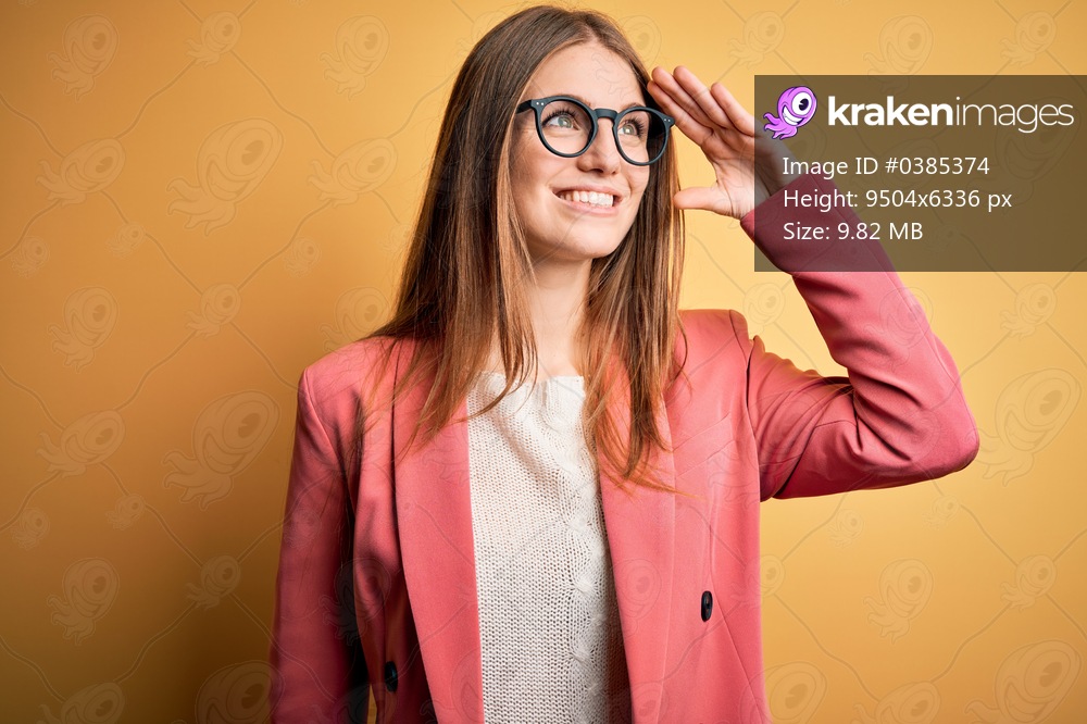 Young beautiful redhead woman wearing jacket and glasses over isolated yellow background very happy and smiling looking far away with hand over head. Searching concept.