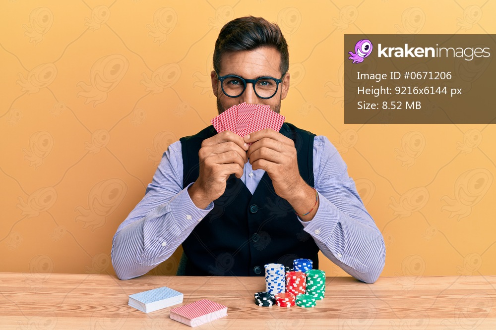 Handsome hispanic croupier man sitting on the table with poker chips and cards smiling with a happy and cool smile on face. showing teeth. 