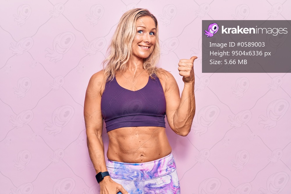 Middle age caucasian blonde woman wearing sportswear over pink background smiling with happy face looking and pointing to the side with thumb up. 
