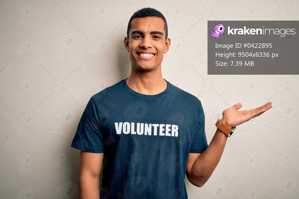 Young handsome african american man volunteering wearing t-shirt with volunteer message smiling cheerful presenting and pointing with palm of hand looking at the camera.