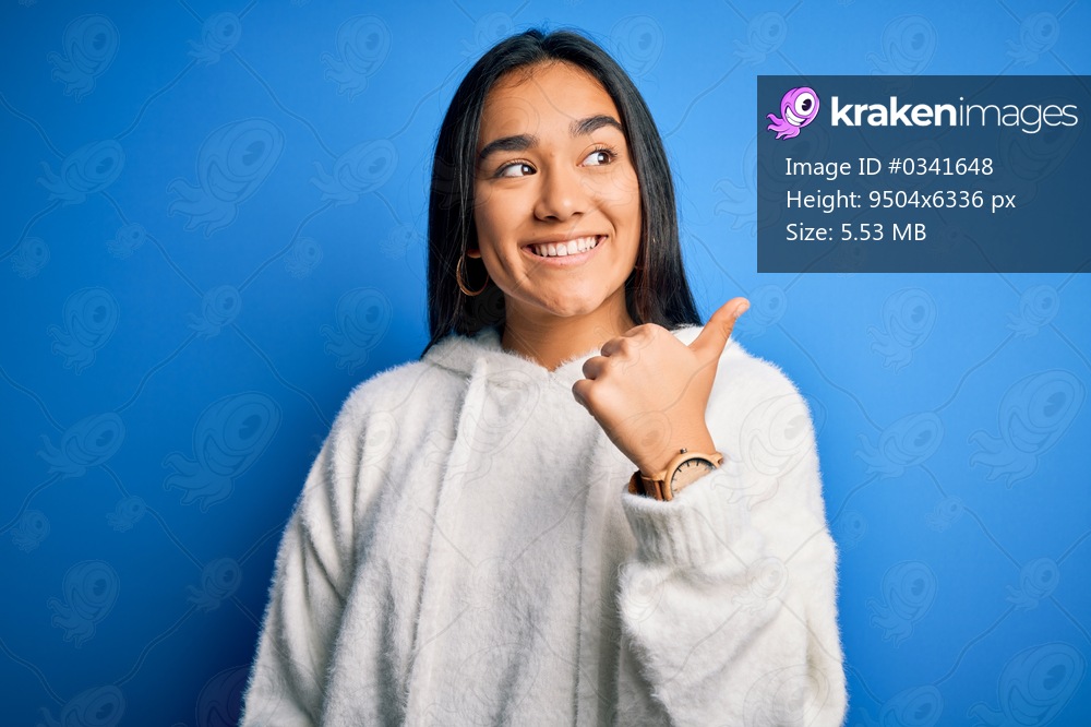 Young beautiful asian sportswoman wearing sweatshirt standing over isolated blue background smiling with happy face looking and pointing to the side with thumb up.