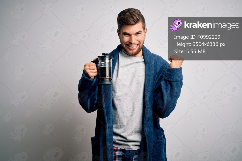 Young blond man with beard and blue eyes wearing pajama making coffe using coffeemaker screaming proud and celebrating victory and success very excited, cheering emotion