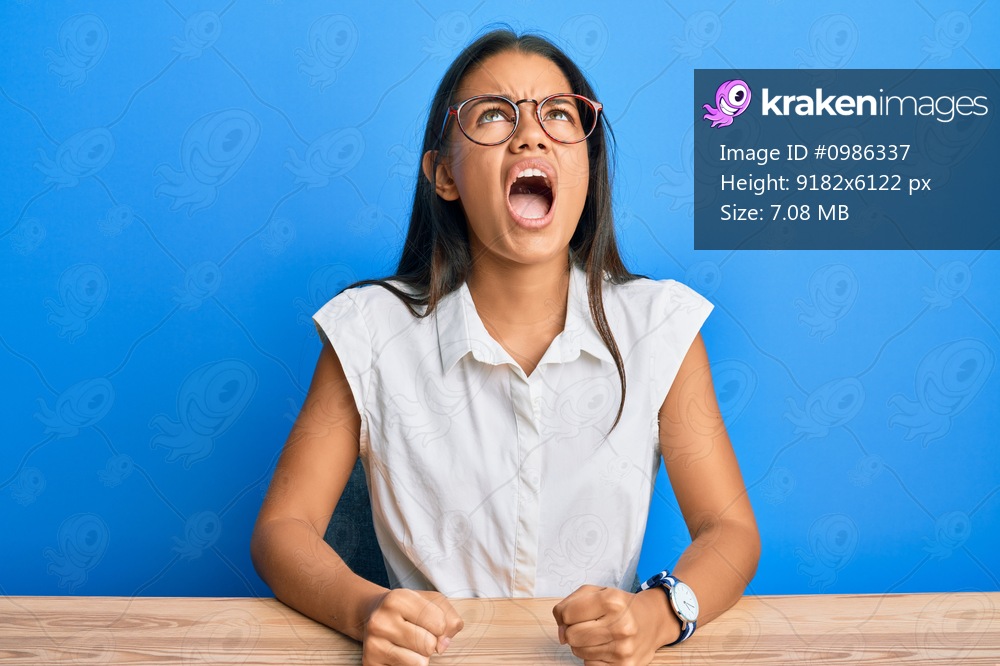 Beautiful hispanic woman wearing casual clothes sitting on the table angry and mad screaming frustrated and furious, shouting with anger. rage and aggressive concept. 