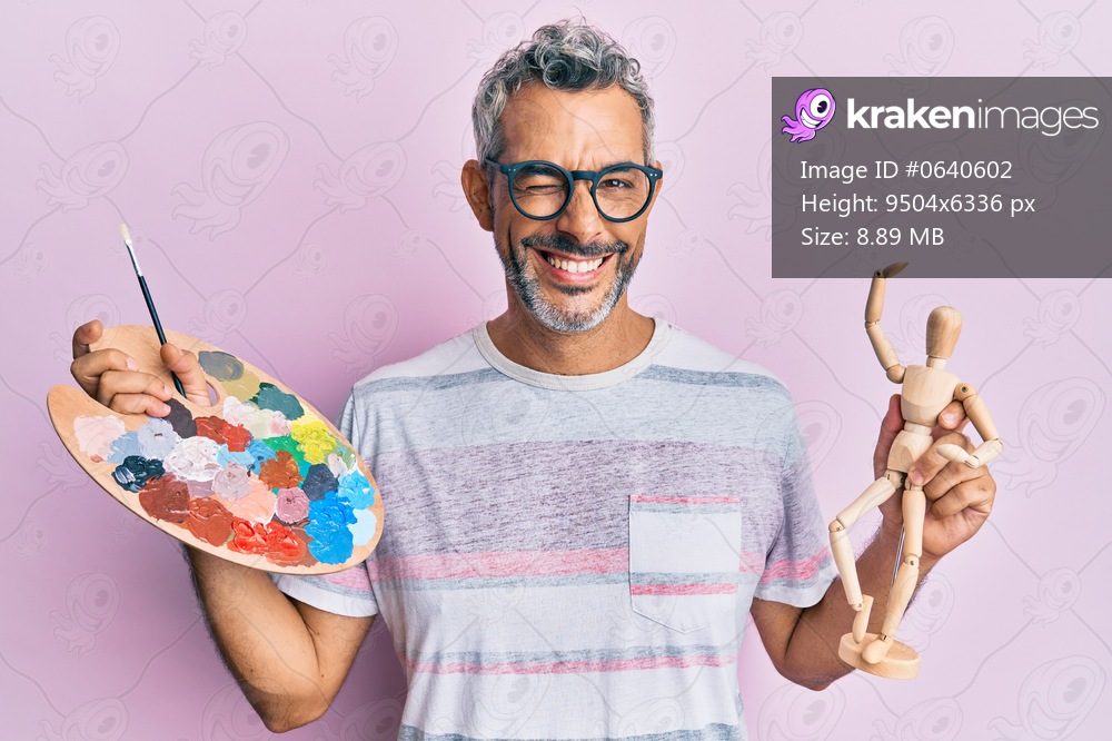 Middle age grey-haired man holding palette and manikin winking looking at the camera with sexy expression, cheerful and happy face. 