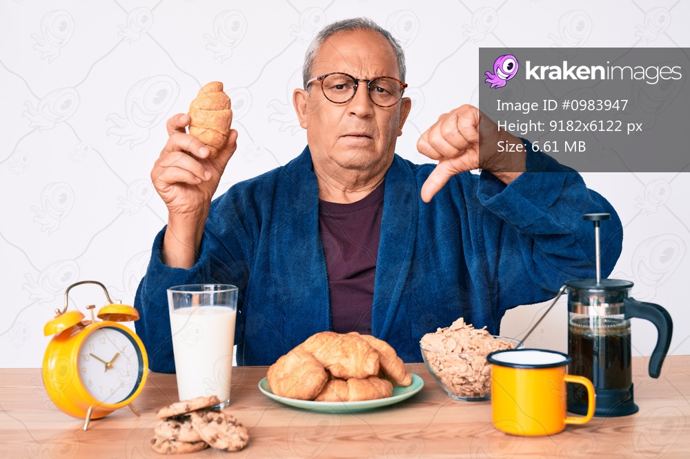 Senior handsome man with gray hair sitting on the table eating croissant for breakfast with angry face, negative sign showing dislike with thumbs down, rejection concept 