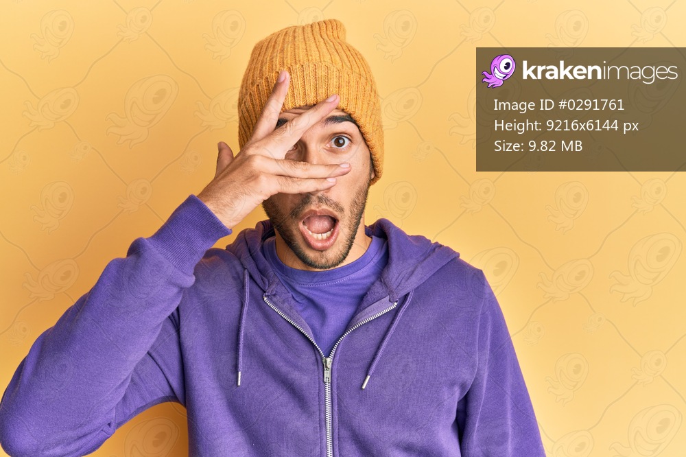 Young handsome man wearing wool winter hat peeking in shock covering face and eyes with hand, looking through fingers afraid 