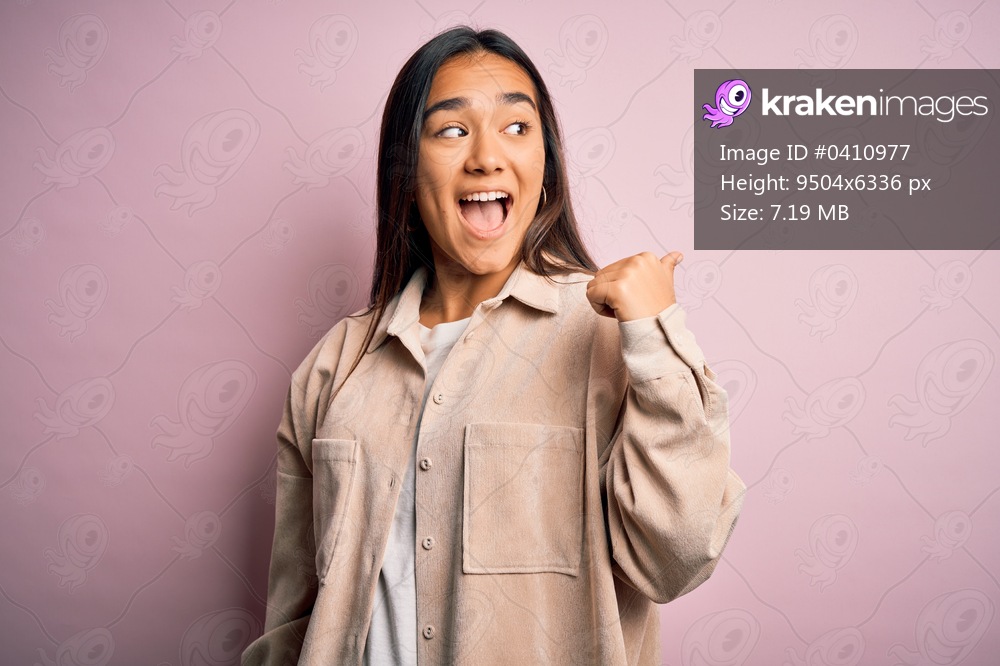 Young beautiful asian woman wearing casual shirt standing over pink background smiling with happy face looking and pointing to the side with thumb up.