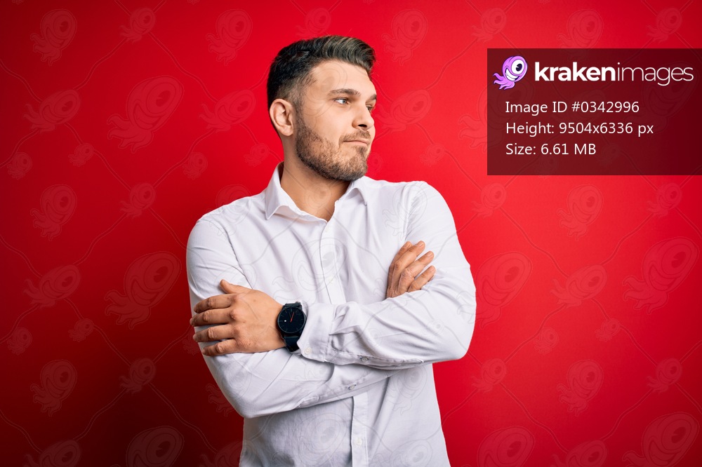 Young business man with blue eyes wearing elegant shirt standing over red isolated background looking to the side with arms crossed convinced and confident
