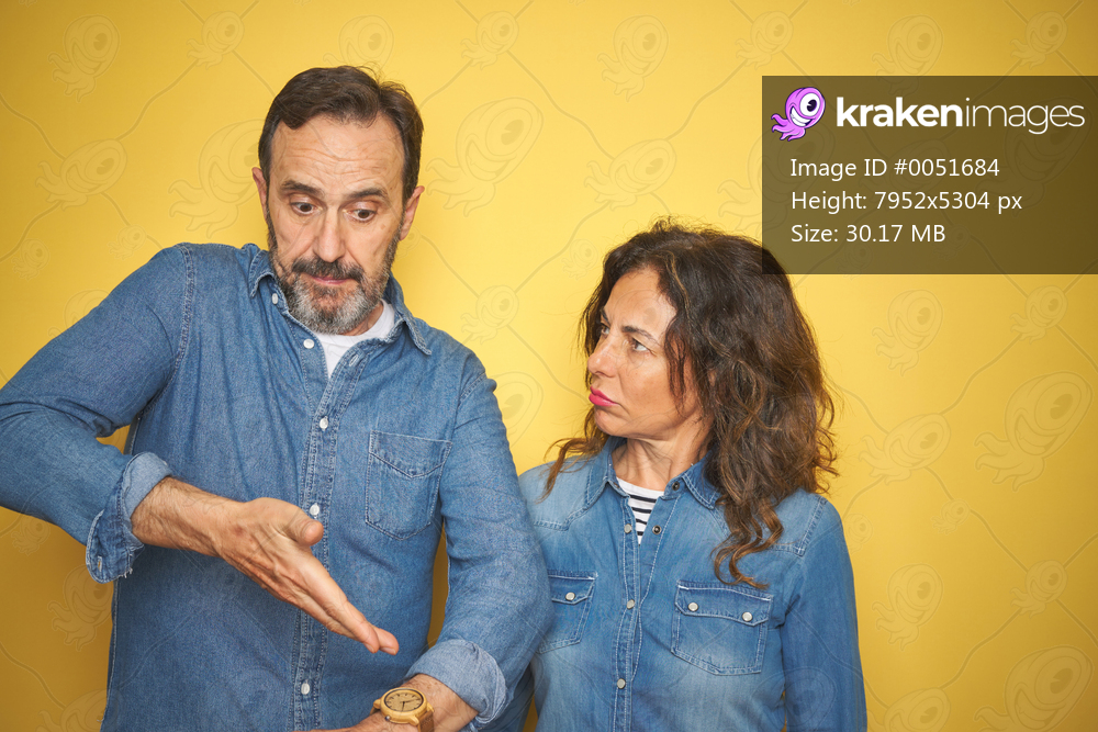 Beautiful middle age couple together standing over isolated yellow background In hurry pointing to watch time, impatience, upset and angry for deadline delay