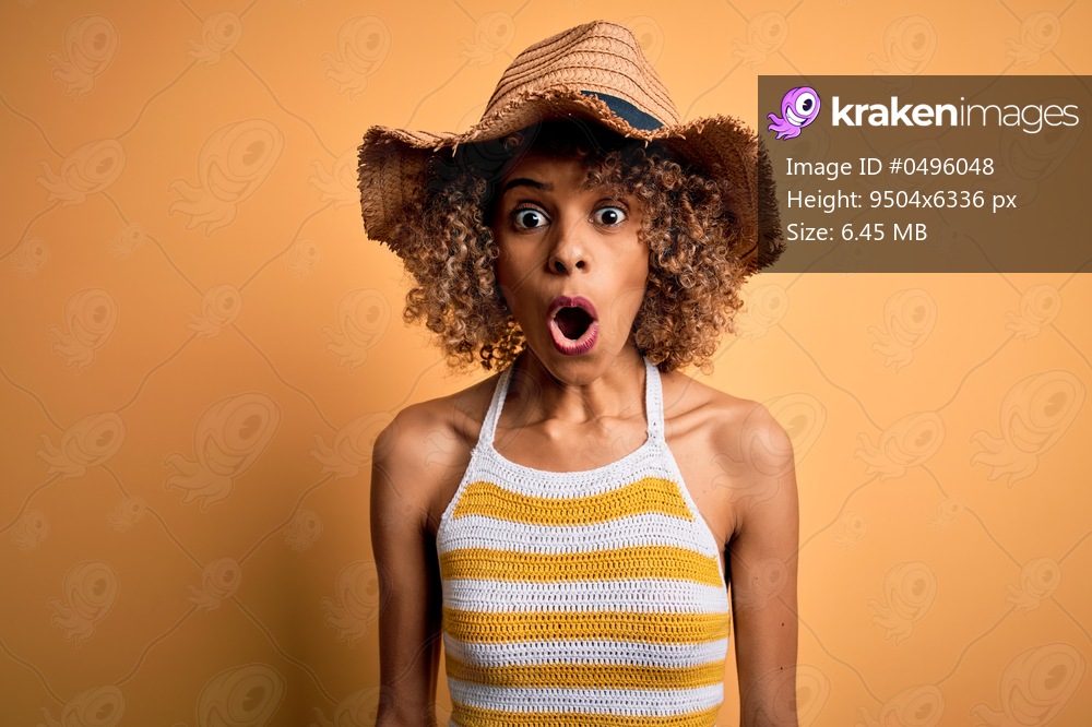 African american tourist woman with curly on vacation wearing summer hat and striped t-shirt afraid and shocked with surprise expression, fear and excited face.
