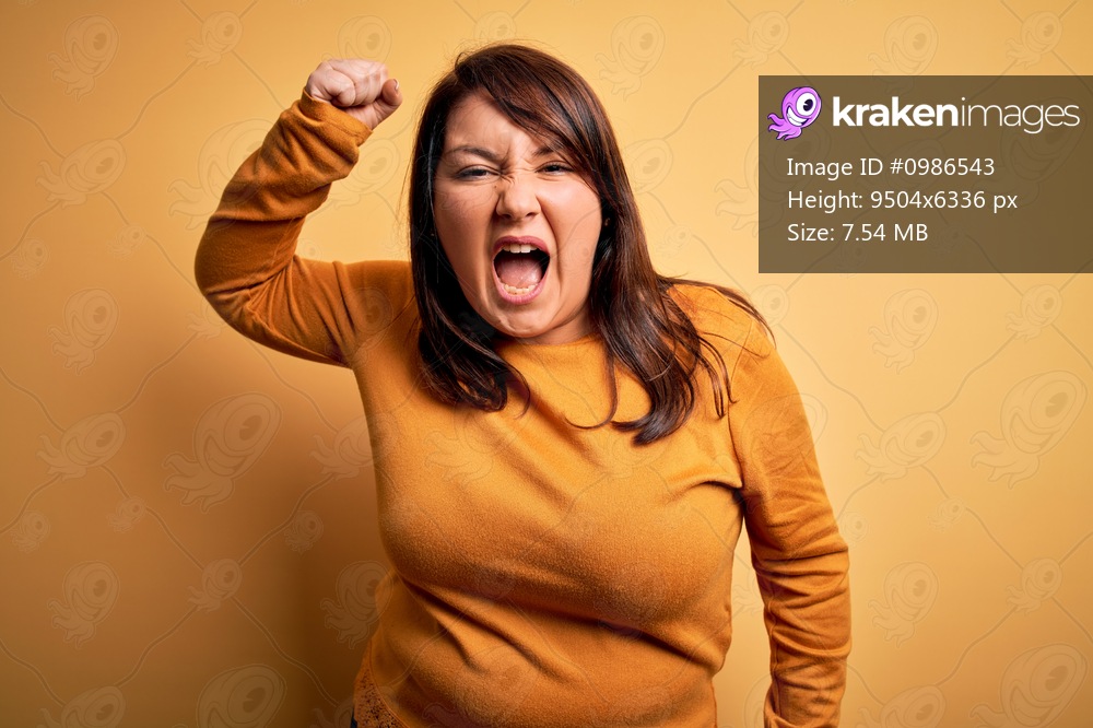 Beautiful brunette plus size woman wearing casual sweater over isolated yellow background angry and mad raising fist frustrated and furious while shouting with anger. Rage and aggressive concept.