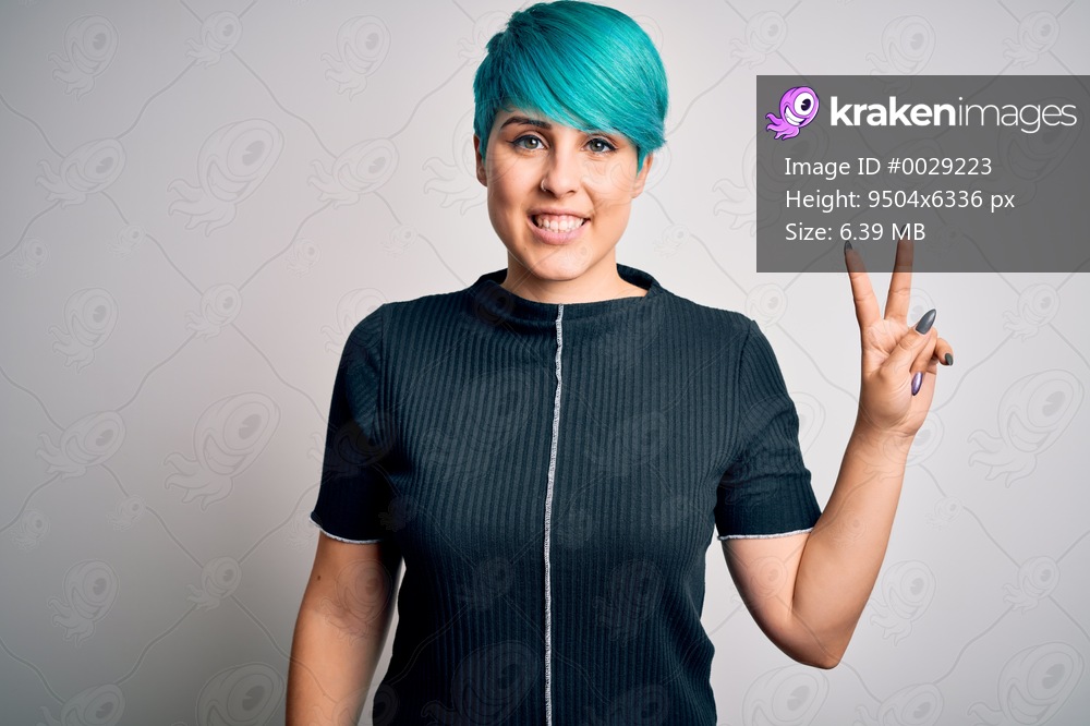 Young beautiful woman with blue fashion hair wearing casual t-shirt over white background smiling with happy face winking at the camera doing victory sign. Number two.