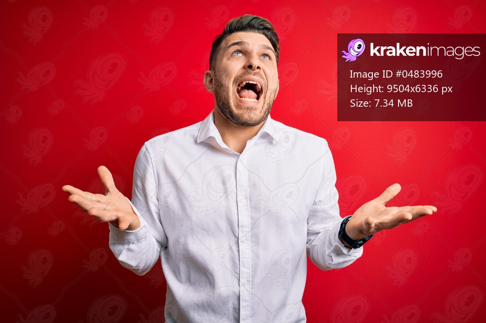 Young business man with blue eyes wearing elegant shirt standing over red isolated background crazy and mad shouting and yelling with aggressive expression and arms raised. Frustration concept.