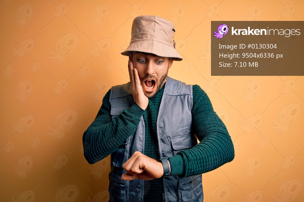 Handsome tourist man with beard on vacation wearing explorer hat over yellow background Looking at the watch time worried, afraid of getting late