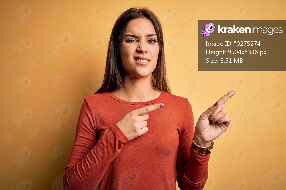 Young beautiful brunette woman wearing casual t-shirt standing over yellow background Pointing aside worried and nervous with both hands, concerned and surprised expression
