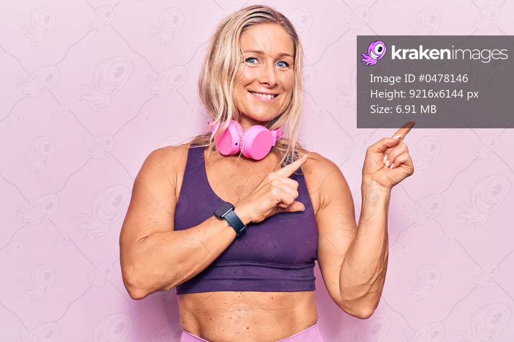 Middle age caucasian blonde woman wearing gym clothes and using headphones smiling and looking at the camera pointing with two hands and fingers to the side. 
