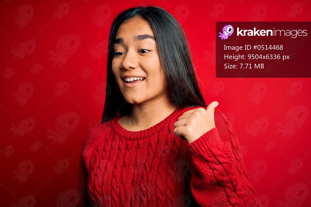 Young beautiful asian woman wearing casual sweater standing over isolated red background smiling with happy face looking and pointing to the side with thumb up.